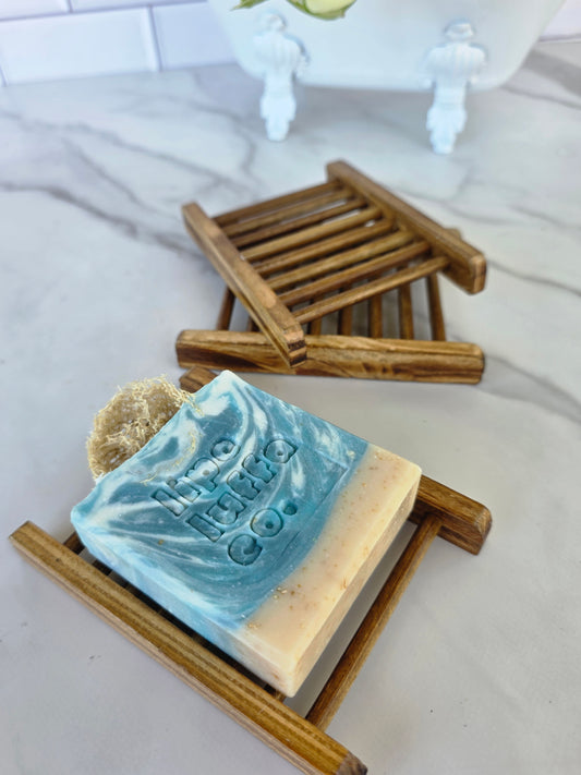 A handmade bar soap bar with a blue and cream swirl pattern and a natural luffa on top, displayed on a small wooden soap dish. A second soap dish is visible in the background on a marble surface.