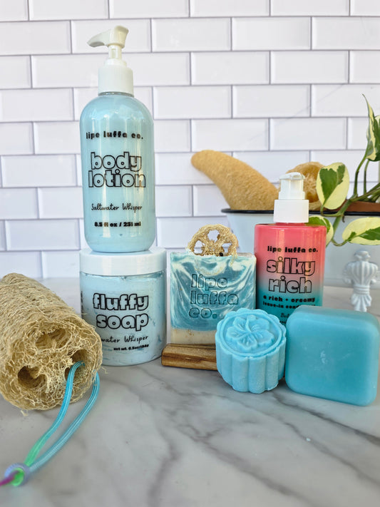 A collection of Lipe Luffa co. bath and body products arranged on a marble countertop against a white tile backsplash.